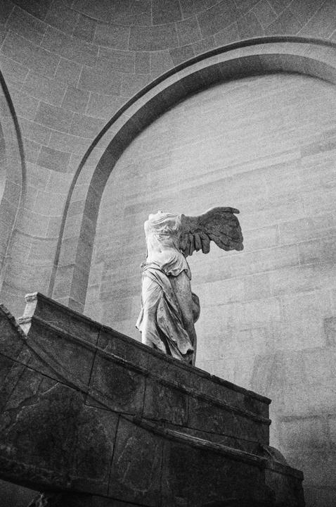 A headless winged statue stands on a pedestal in a large room with arched ceilings.