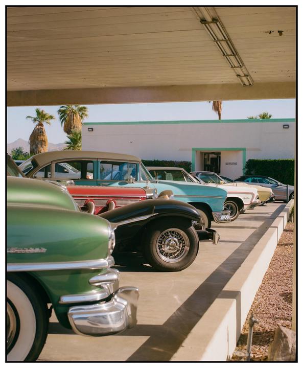 A line of vintage cars is parked under a covered structure with palm trees and a building in the background.