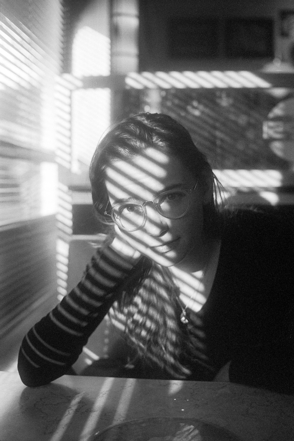 A woman with glasses rests her head on her hand, illuminated by striped shadows from blinds.