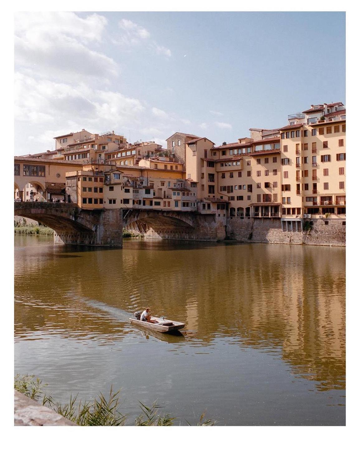 A man in a small boat navigates a river with a historic bridge and buildings lining its banks.