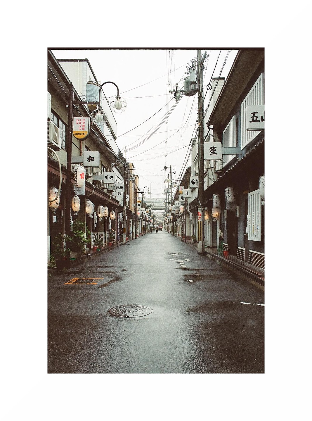 A narrow street in Osaka is lined with buildings and lanterns under a cloudy sky.