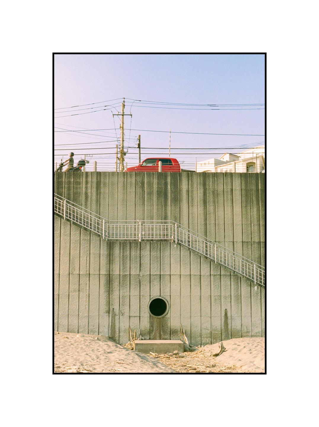 A red car is parked on a concrete wall with a staircase leading down to a sandy area.