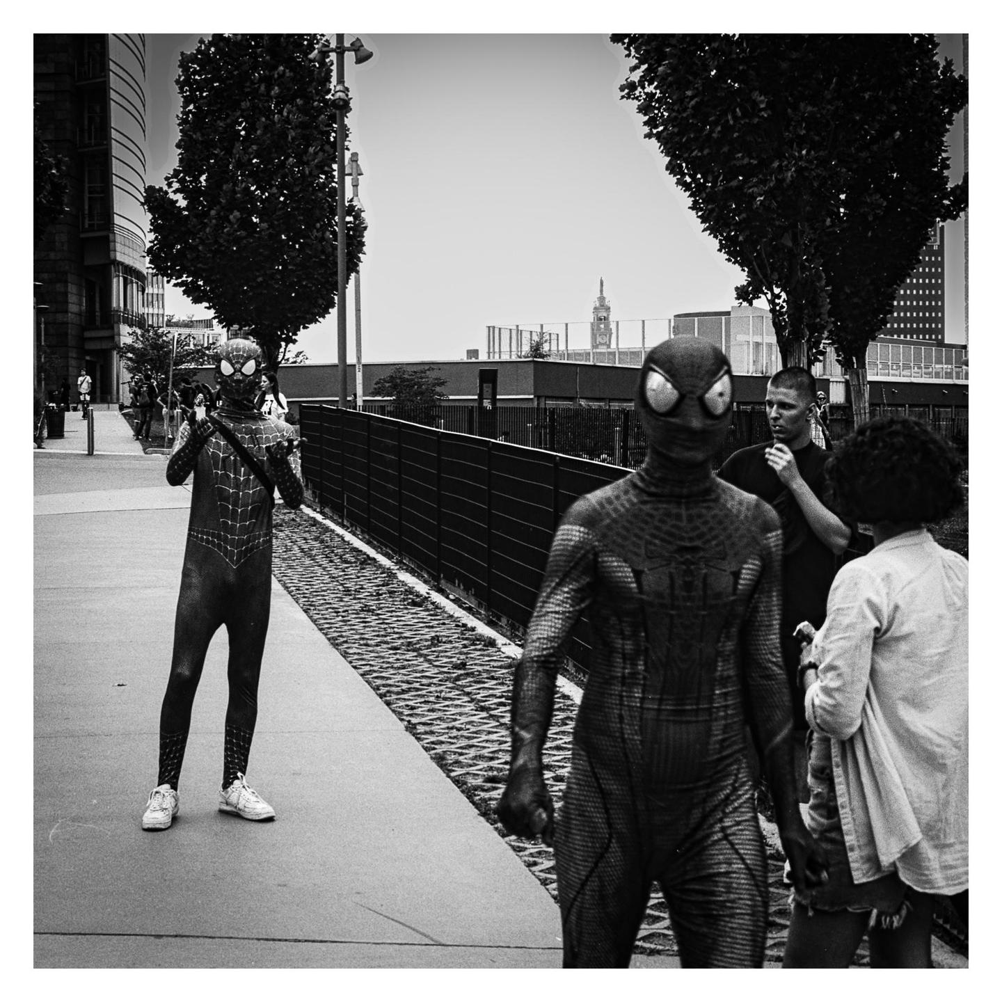 Two people in spider-man costumes stand on a sidewalk with other people and buildings in the background.