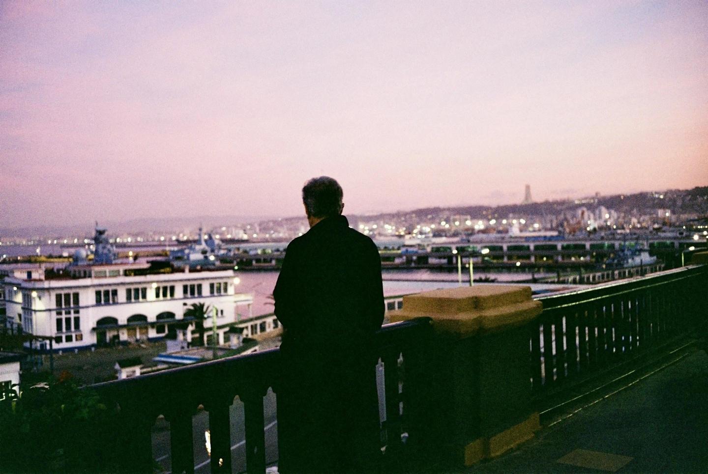 A man stands on a balcony overlooking a city and harbor at dusk.