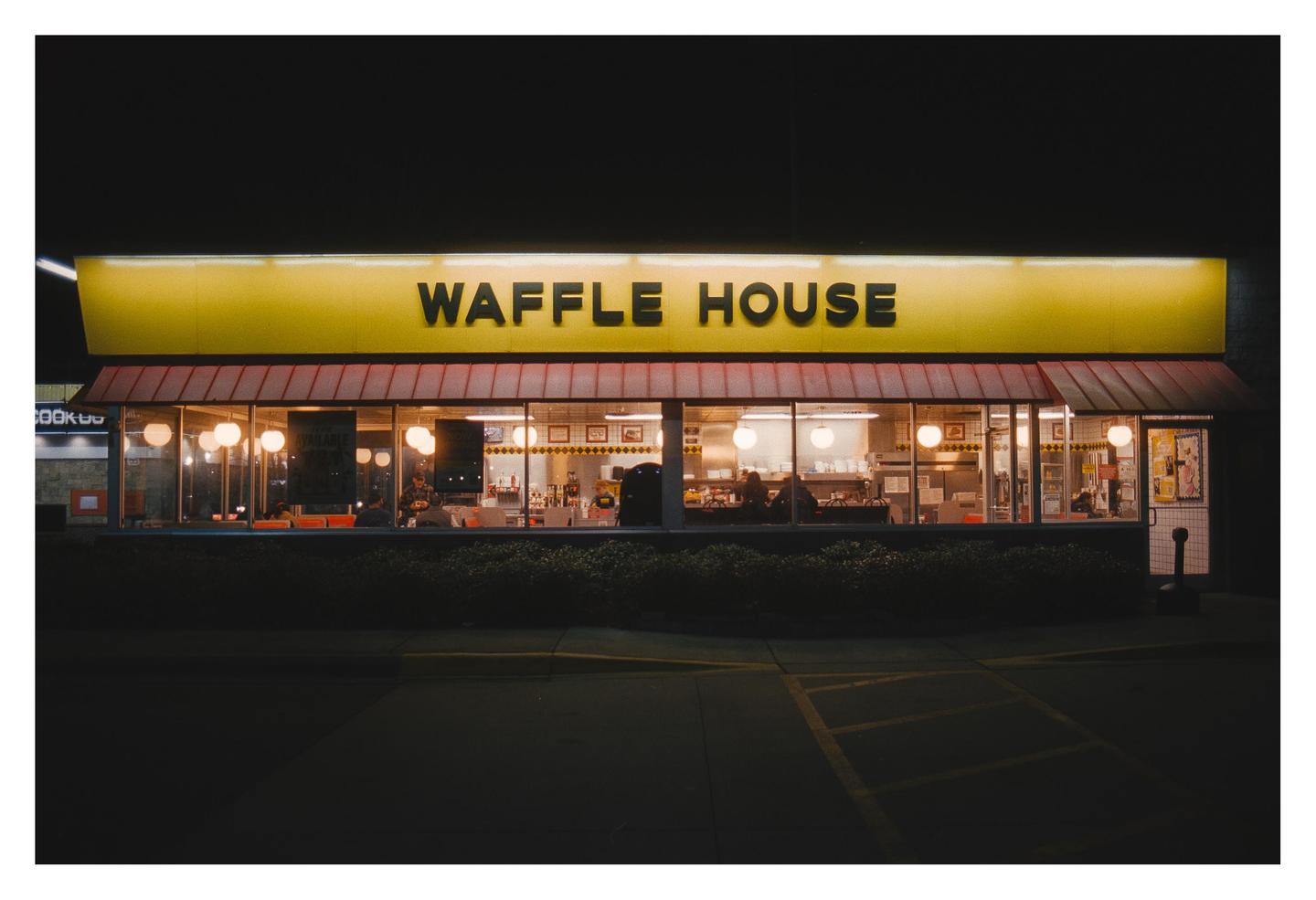 The exterior of a Waffle House restaurant is illuminated at night, with people visible inside through the windows.