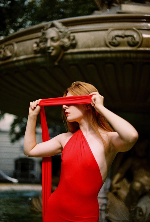 A woman in a red dress holds a red fabric strip over her eyes, standing in front of a stone fountain.
