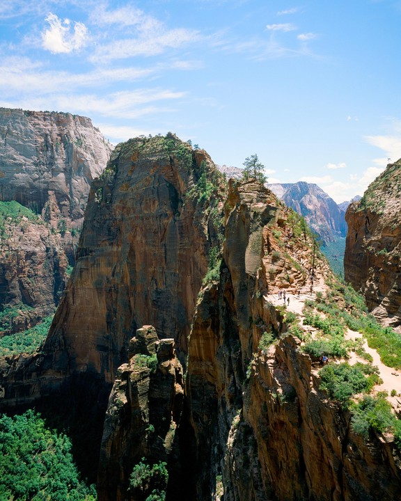 A group of people hike along a narrow trail on a steep, rocky mountain ridge under a bright blue sky.