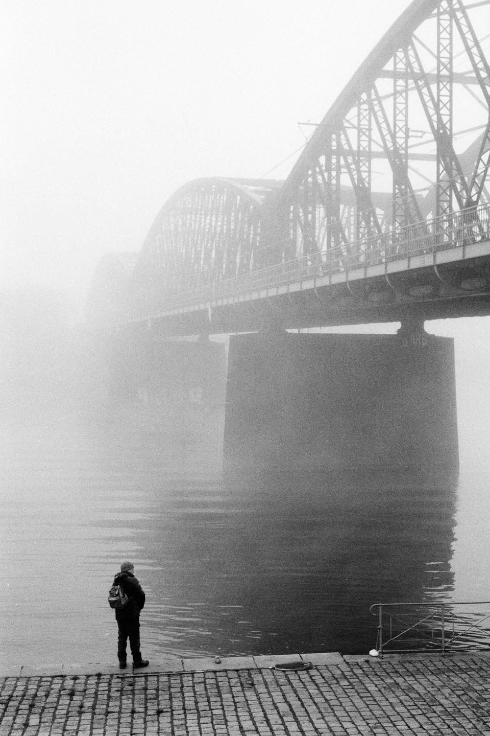 A person stands on a cobblestone riverbank looking out at a bridge shrouded in fog.