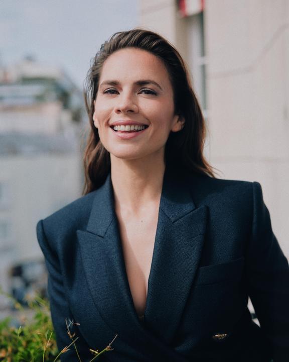 A woman with long hair smiles at the camera while wearing a dark blazer outdoors.