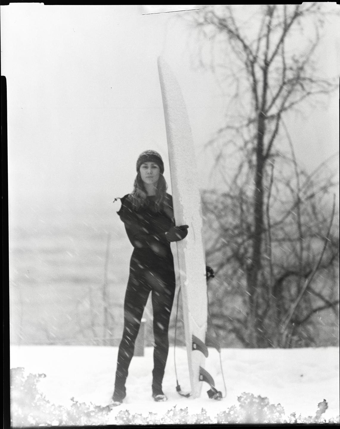 A woman in a wetsuit and beanie stands in the snow next to a surfboard.