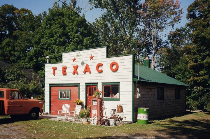 An old Texaco gas station with an orange truck parked outside is surrounded by trees.