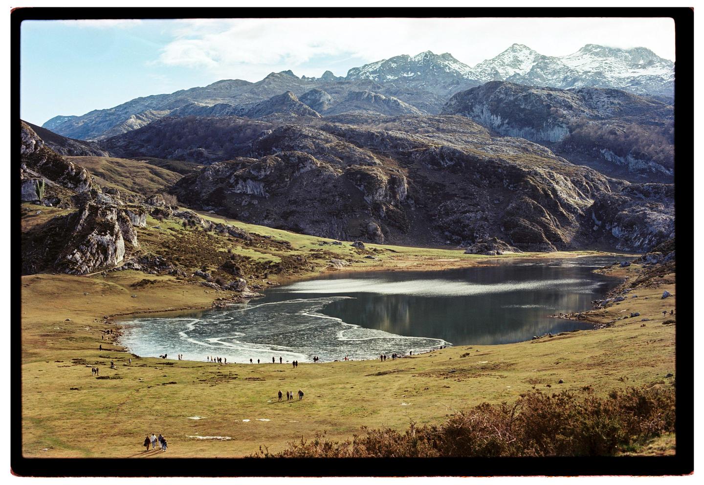 A partially frozen lake is surrounded by grassy hills and snow-capped mountains under a bright sky.