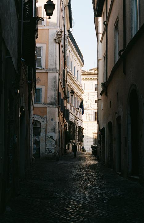 A person walks down a narrow, shadowed cobblestone alley lined with tall buildings.