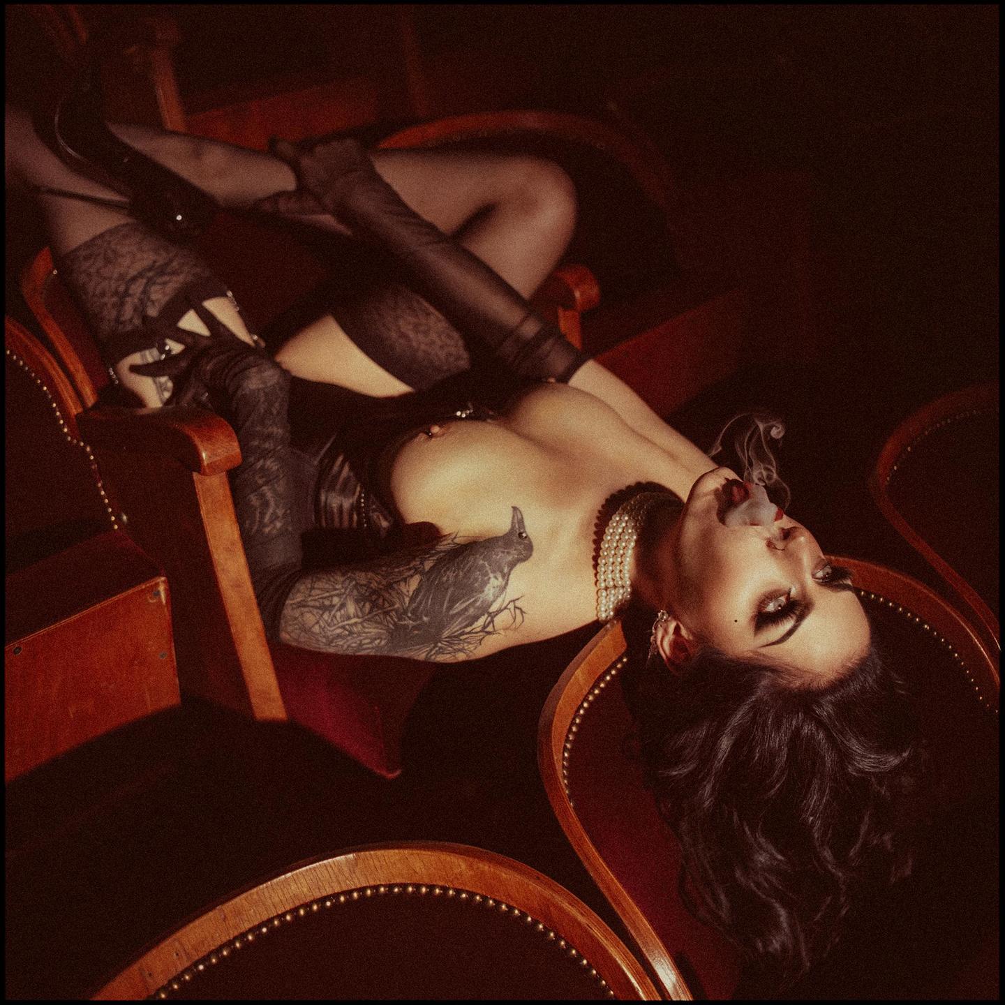 A woman with a tattoo on her arm is lying on a chair, exhaling smoke from her mouth.