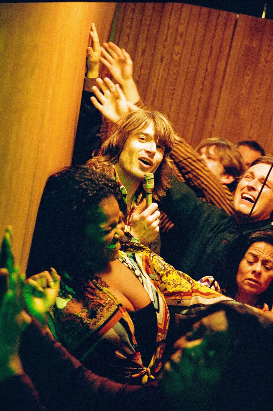 A man with long hair sings into a microphone while surrounded by other people, some of whom are raising their hands.