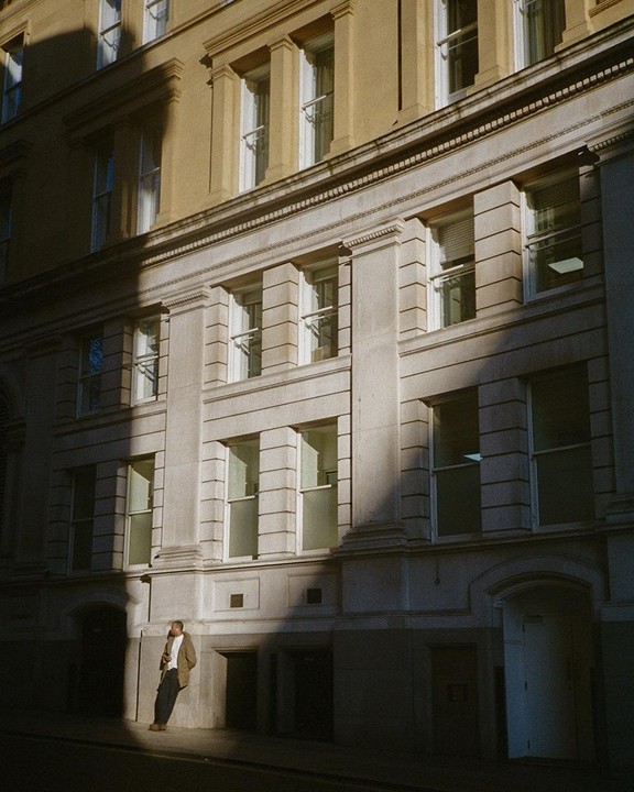 A man leans against a stone building with many windows, partially illuminated by sunlight.
