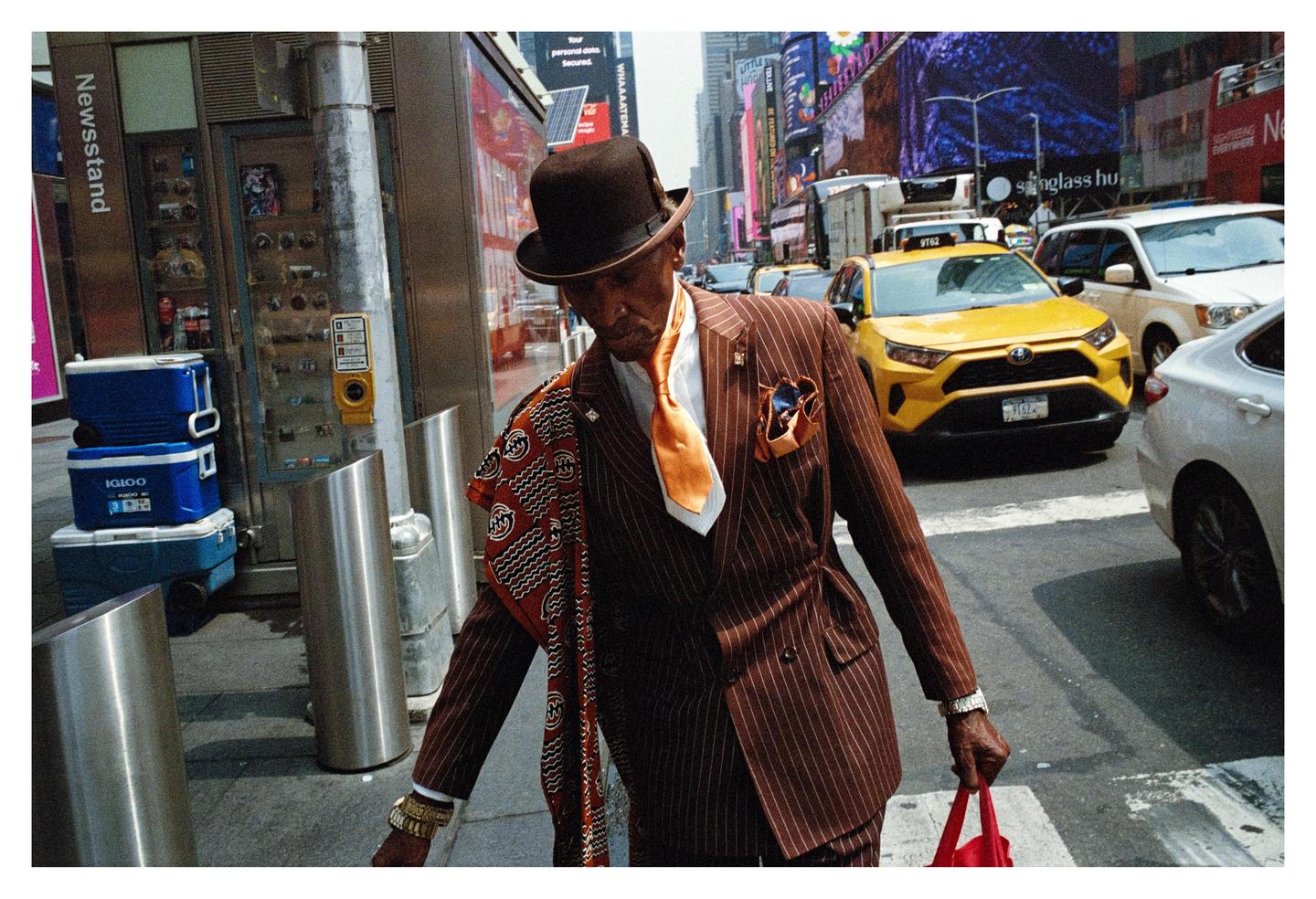 A man in a pinstripe suit and bowler hat walks past a newsstand and yellow taxi in New York.