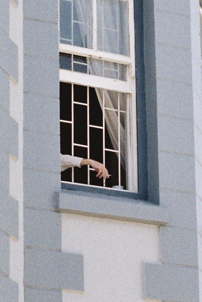 A hand holding a cigarette extends from a window with white bars and sheer curtains.