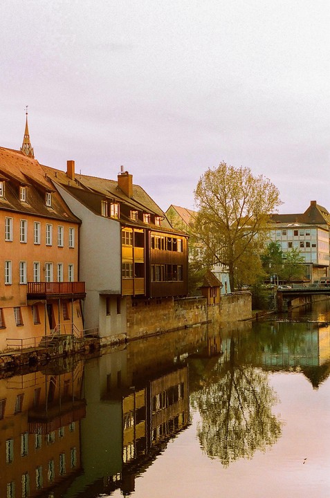 Buildings with orange roofs and a tall tree are reflected in a calm river under a pale sky.