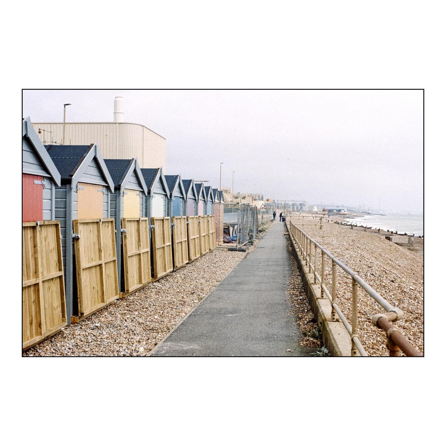 A row of colorful beach huts stands beside a paved path and a pebble beach with a railing.