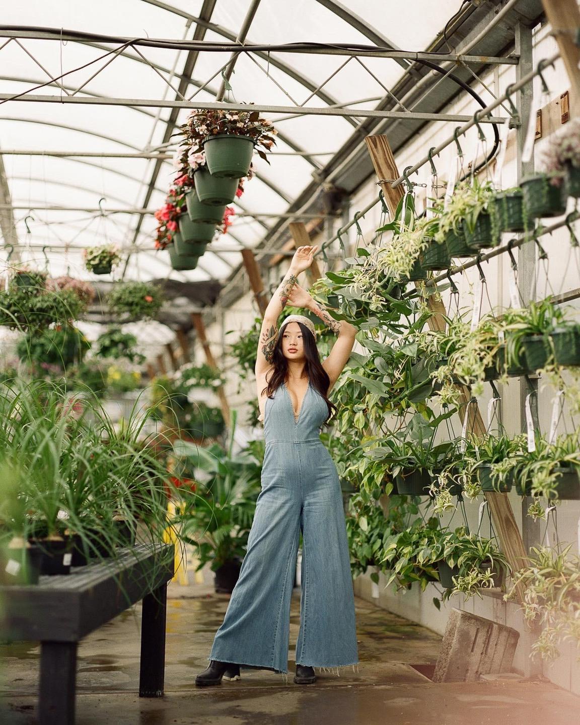 A woman in a denim jumpsuit stands with her arms raised in a greenhouse filled with plants.