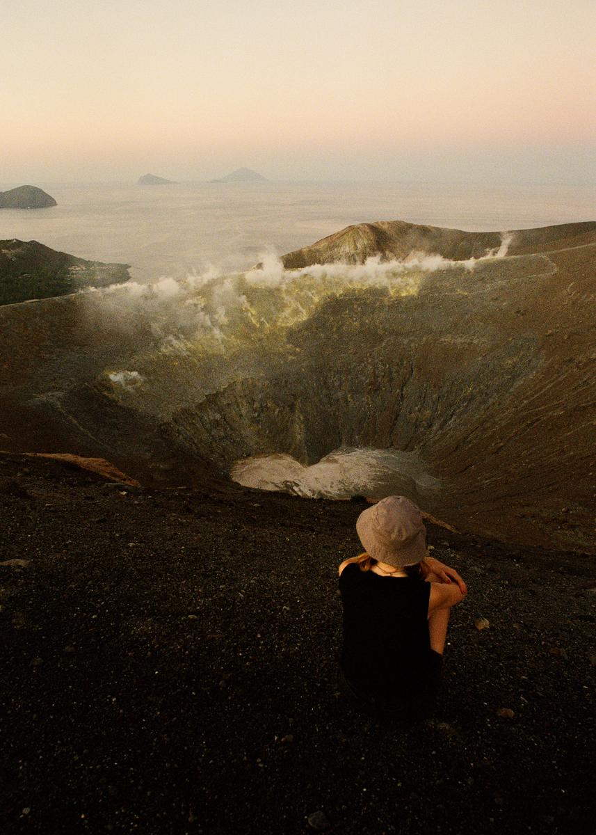 A person sits on the edge of a volcanic crater, looking out at the sea and distant islands at dusk.