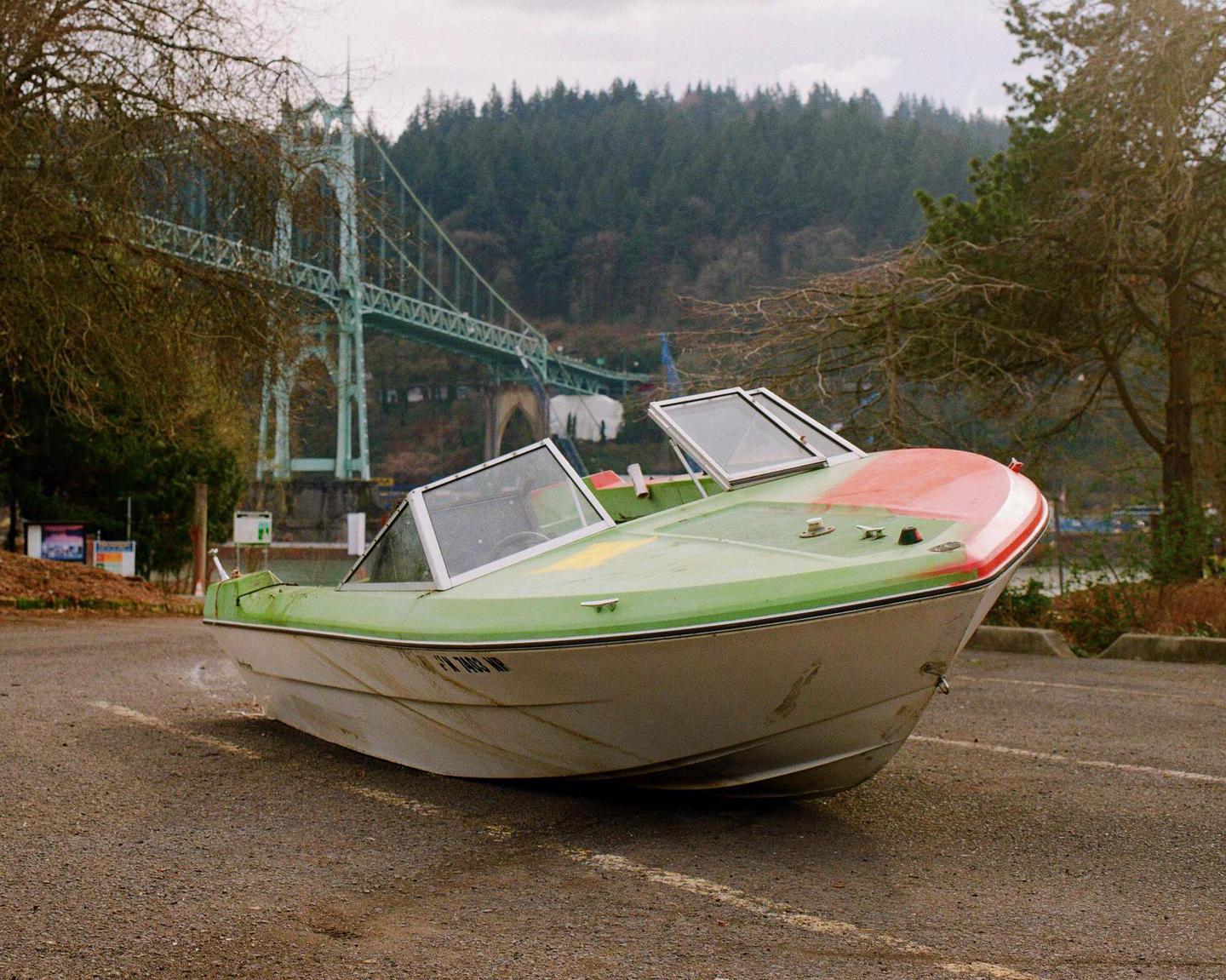 A green and white boat sits on a paved surface with a large bridge and trees in the background.