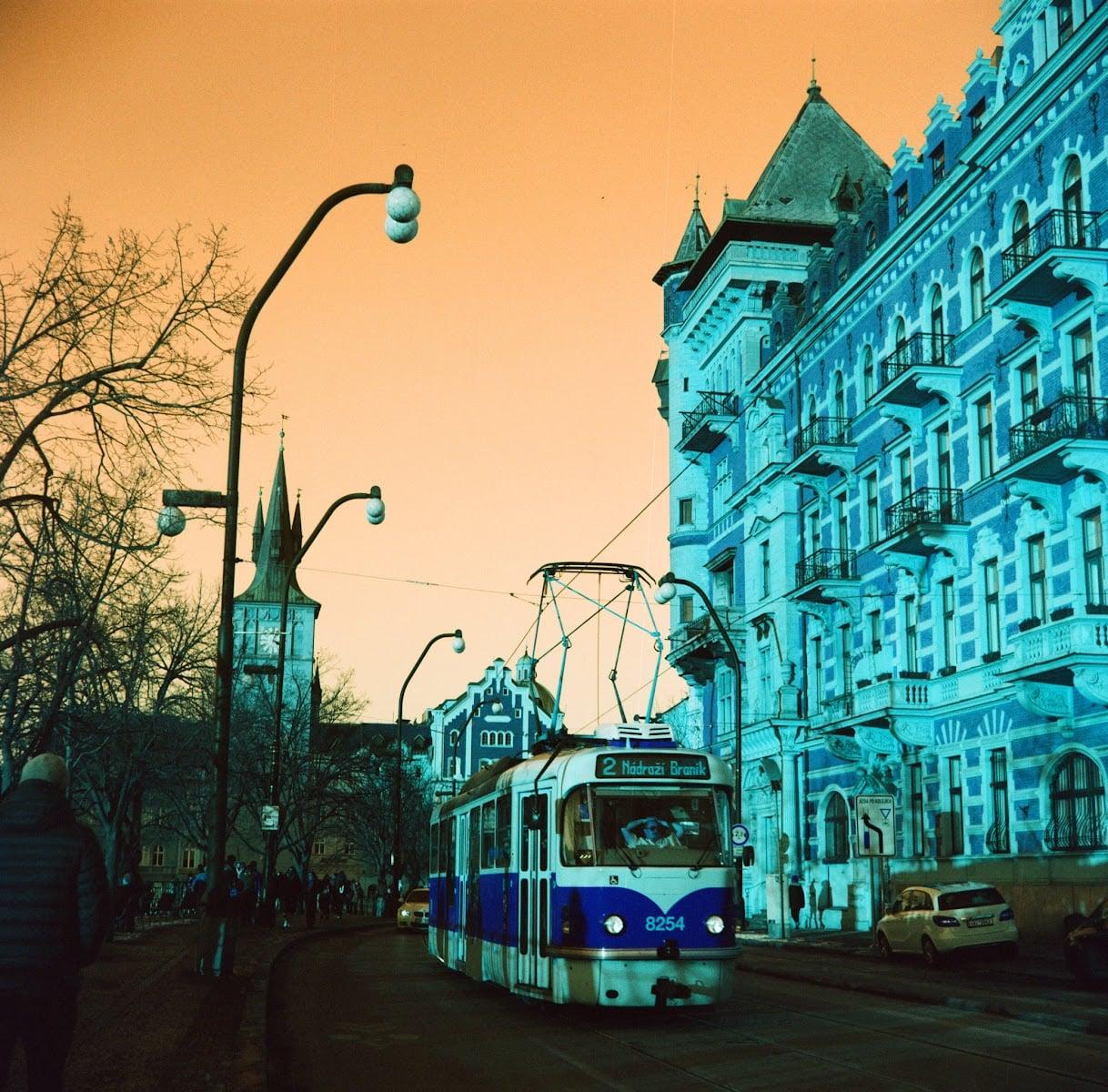A blue and white tram travels down a street lined with ornate blue buildings and bare trees under an orange sky.