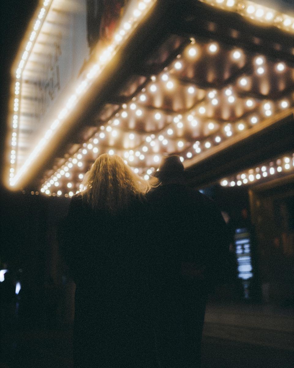 Two people stand with their backs to the viewer, looking up at a brightly lit marquee at night.