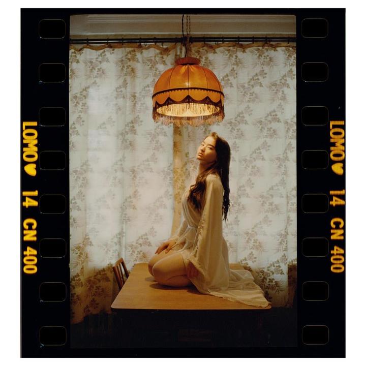 A woman kneels on a wooden table under a hanging lamp, with patterned curtains in the background.