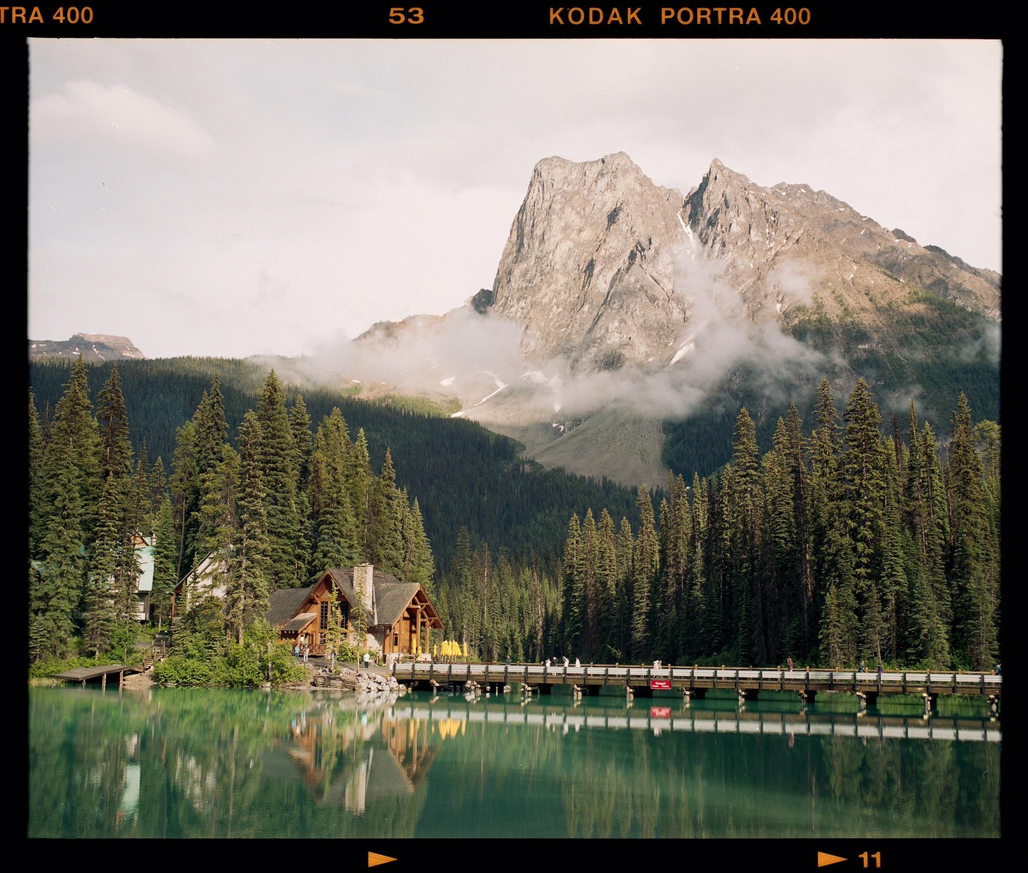 A wooden lodge and a long bridge are reflected in a turquoise lake surrounded by a dense forest and mountains.