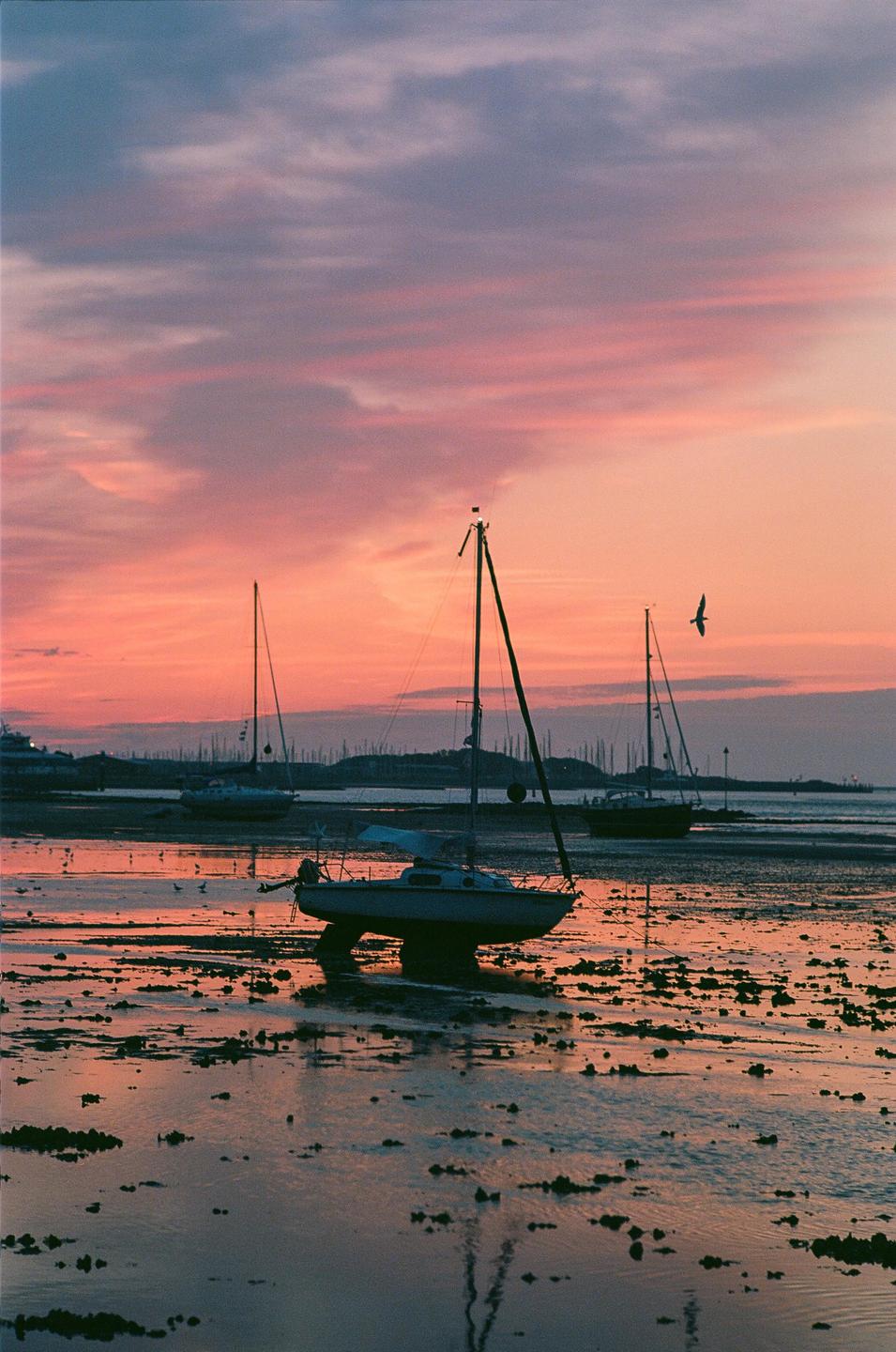 Several sailboats are beached on a muddy shore under a colorful sunset sky.