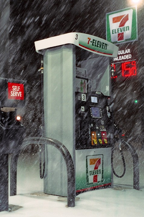 A 7-Eleven gas pump is covered in snow during a winter storm at night.