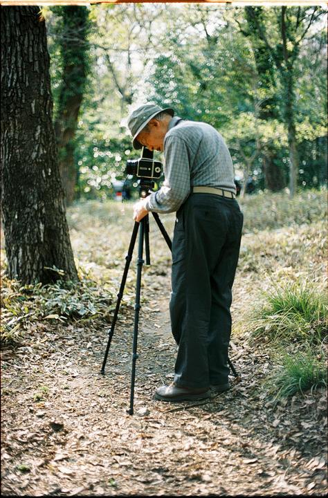 A man wearing a hat is bent over a camera on a tripod in a forest.