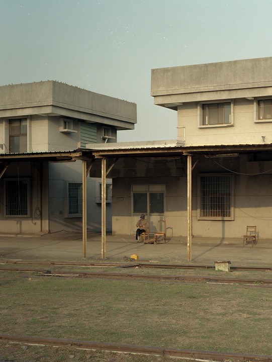 A man sits on a chair under a covered walkway next to a building with train tracks and grass in the foreground.