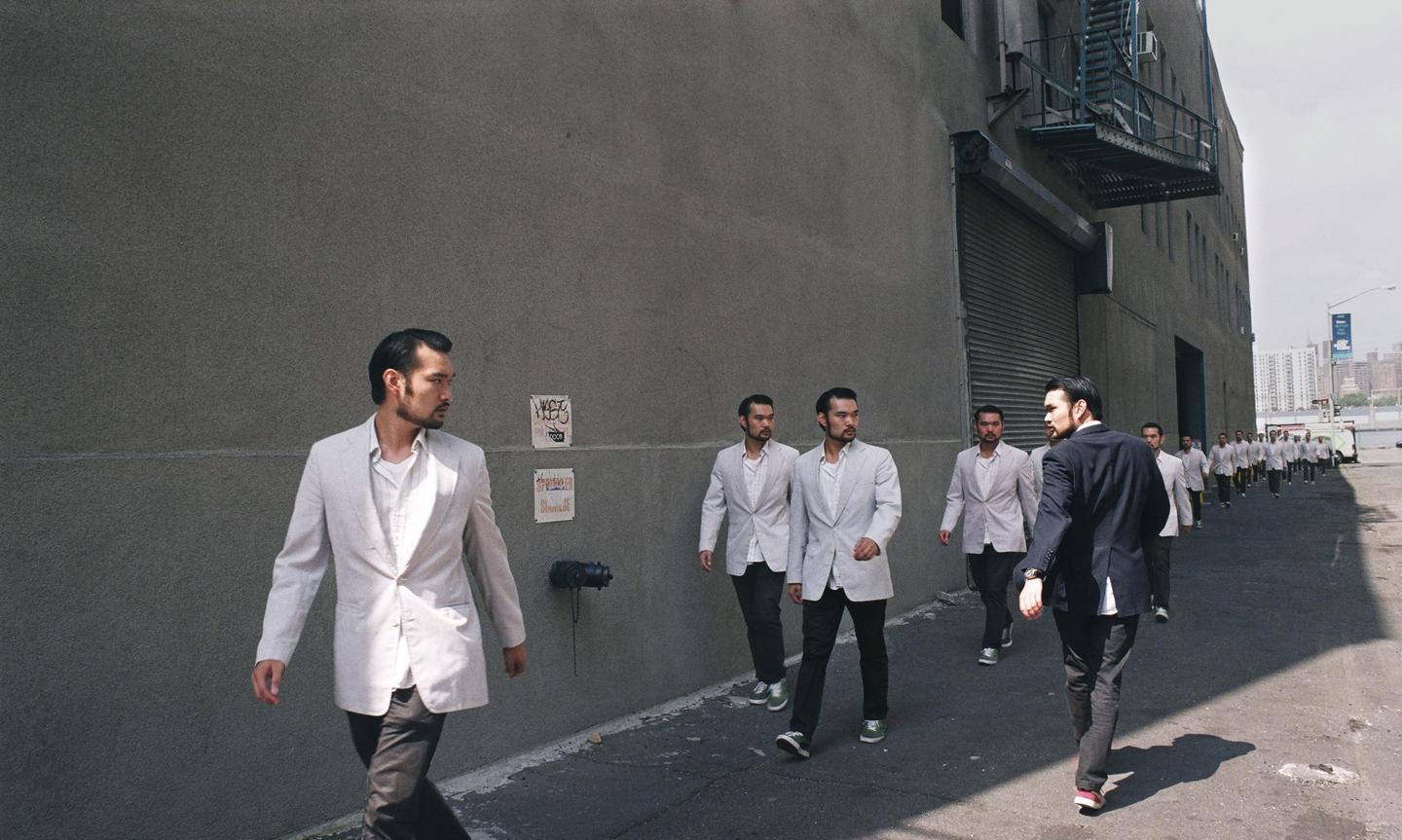 A man in a light jacket walks past a line of identical men in light jackets, with one man in a dark jacket walking away from the viewer.
