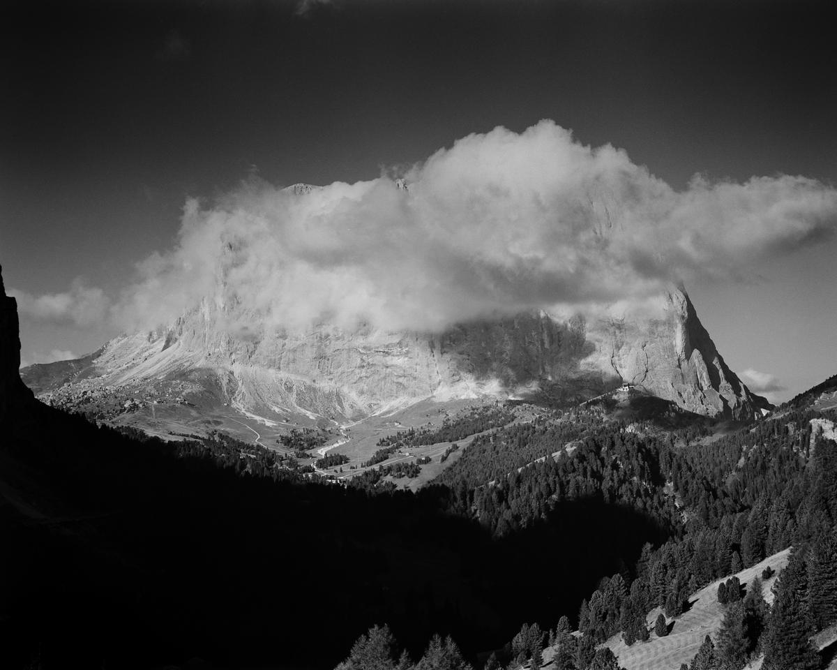 Clouds partially obscure a large mountain peak above a valley with a forest.