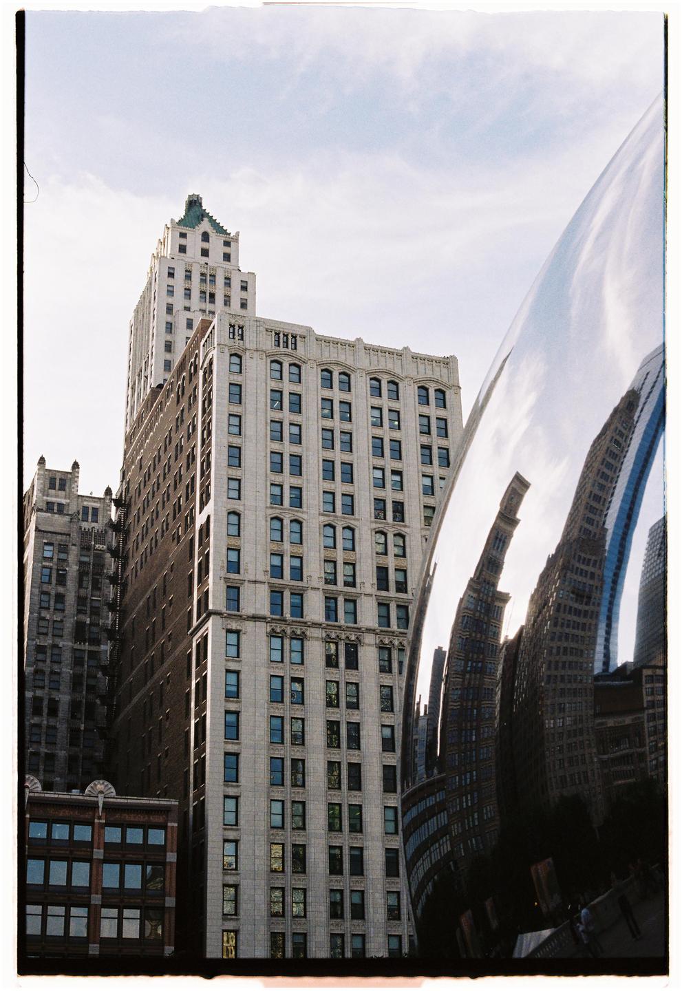 A large, reflective sculpture distorts the reflection of surrounding buildings under a bright sky.