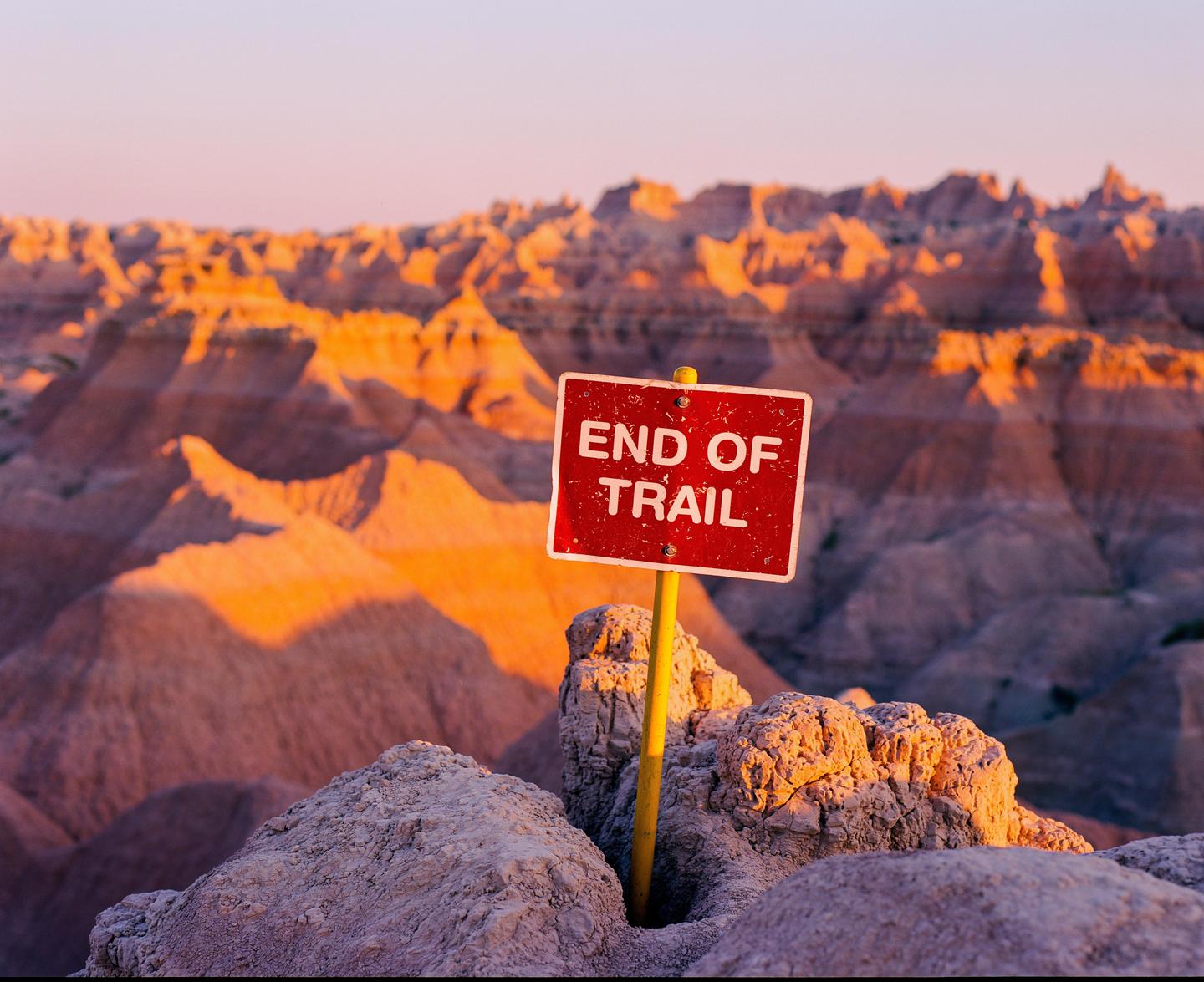 A red sign reading "END OF TRAIL" stands on a rocky outcrop with a badlands landscape in the background during golden hour.