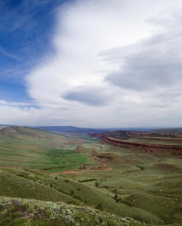 A winding dirt road cuts through a green valley with red rock formations under a partly cloudy sky.