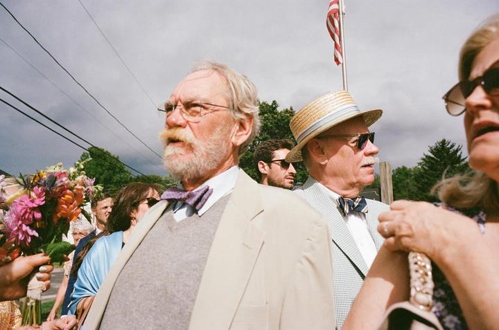 A man with a beard and glasses stands in a crowd of people, with a man in a straw hat and an American flag visible in the background.