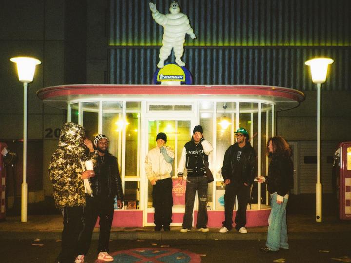 Five men and one woman stand outside a small building with a Michelin Man statue on the roof at night.