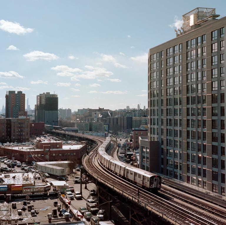 A train travels on elevated tracks through a city with tall buildings and parked cars.