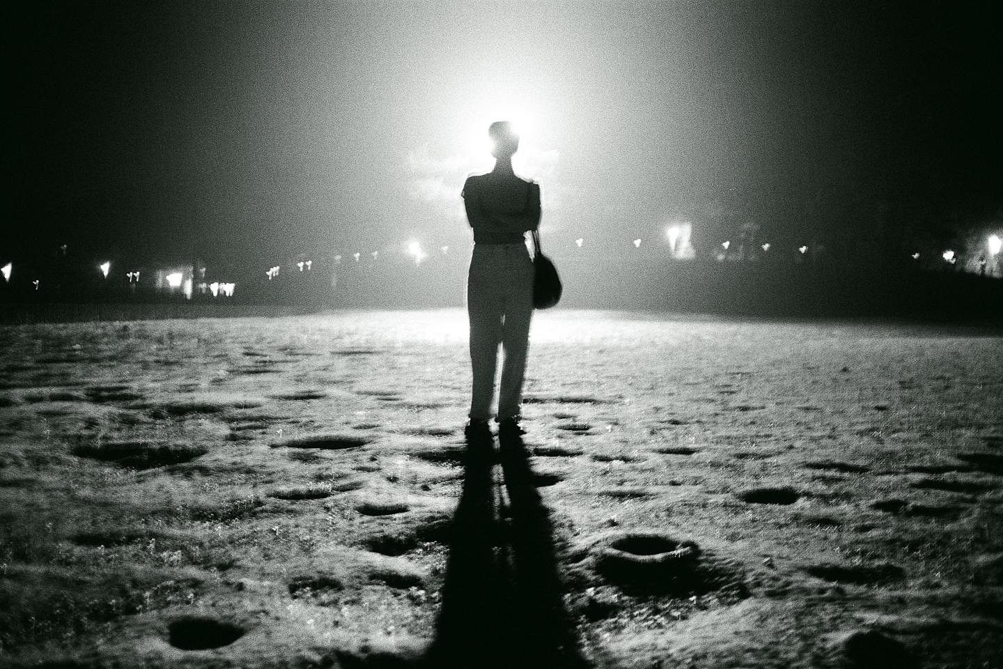 A person stands backlit by a bright light in a dark, textured field with distant lights.