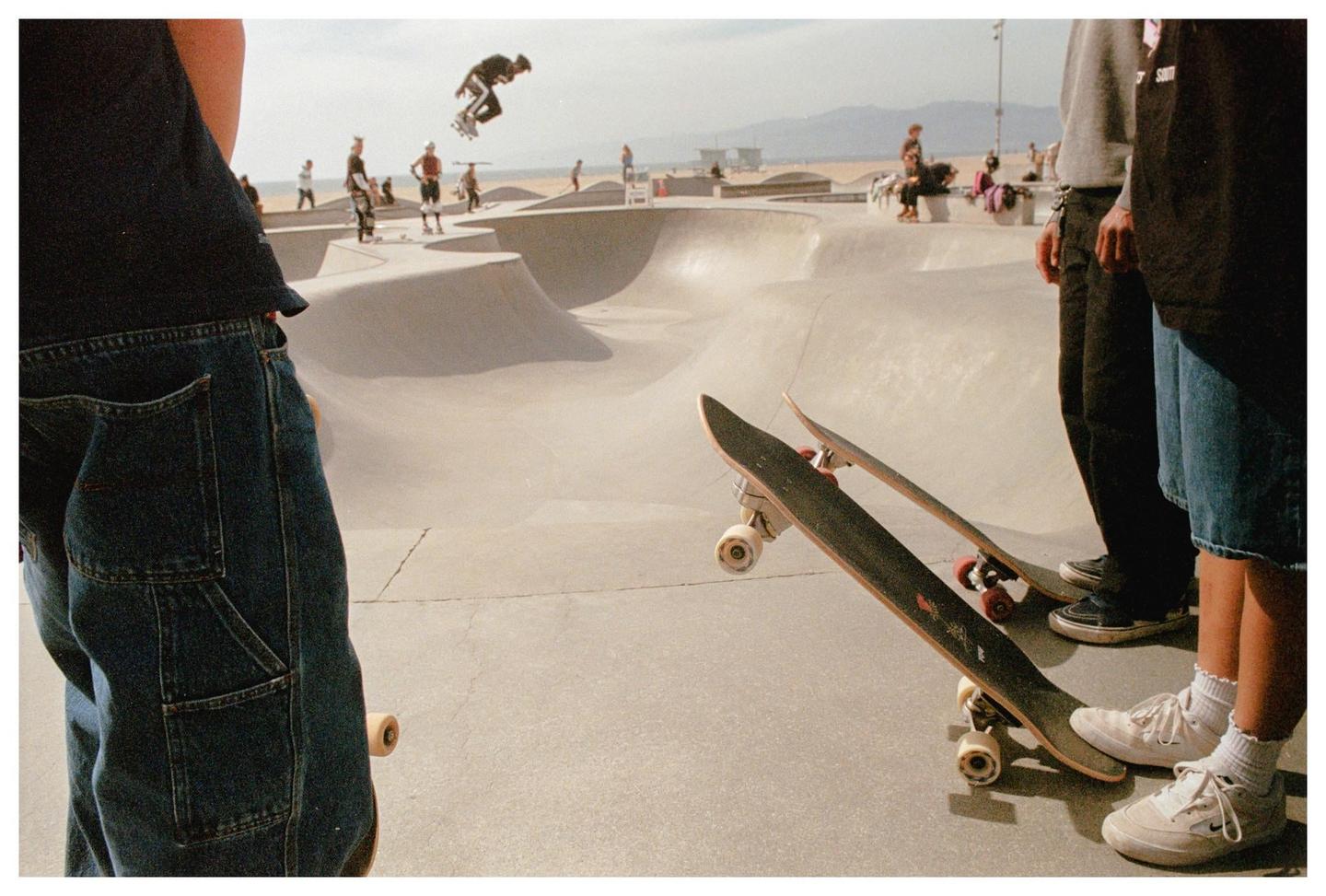 A skateboarder is captured mid-air over a concrete skate park with other people and skateboards visible.