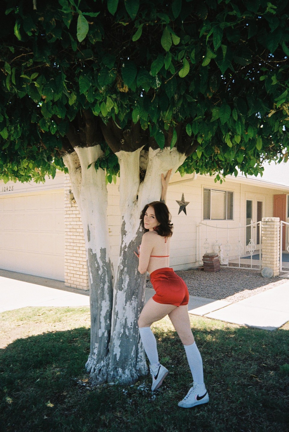 A woman in a red top and shorts leans against a tree with white-painted trunks in front of a house.