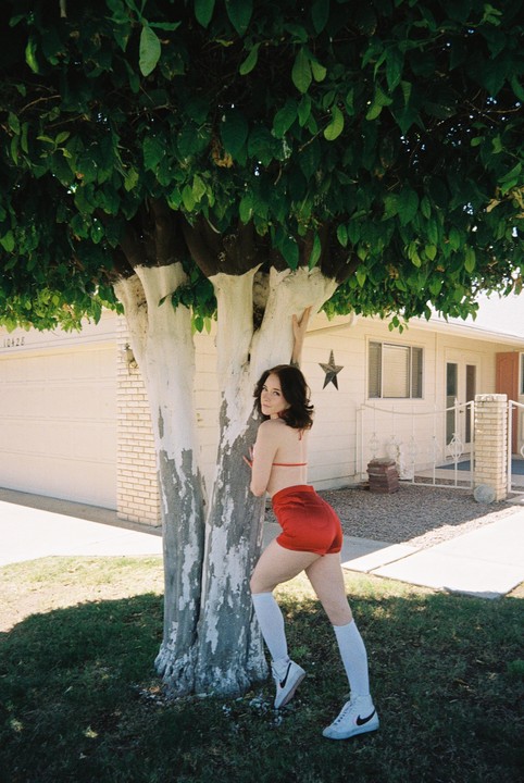 A woman in a red top and shorts leans against a tree with white-painted trunks in front of a house.