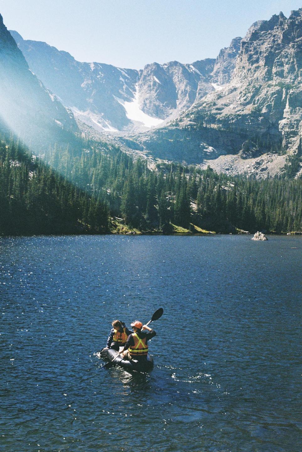 Two people in an inflatable kayak paddle across a lake surrounded by a pine forest and mountains.