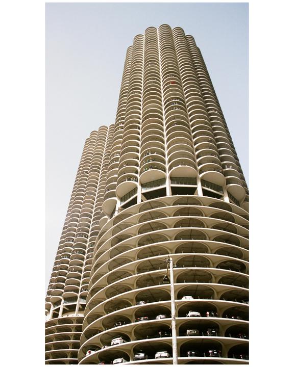 Two tall, cylindrical buildings with many balconies and visible parking levels are shown from a low angle against a light sky.