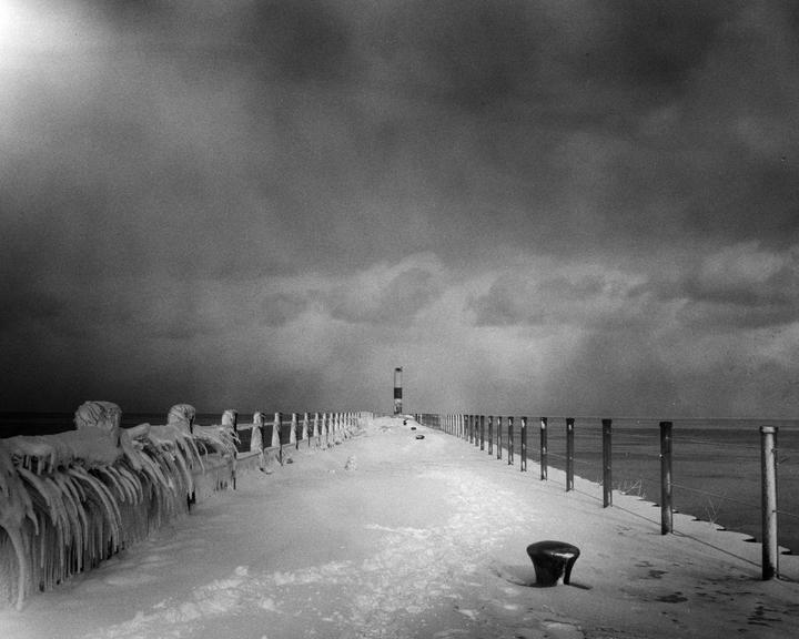 A snow-covered pier with ice formations on a railing leads to a distant lighthouse under a cloudy sky.